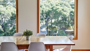 A dining area with a wooden table and white chairs, a potted plant, and large windows offering a view of trees. - Seahaven Village (Barwon Heads)