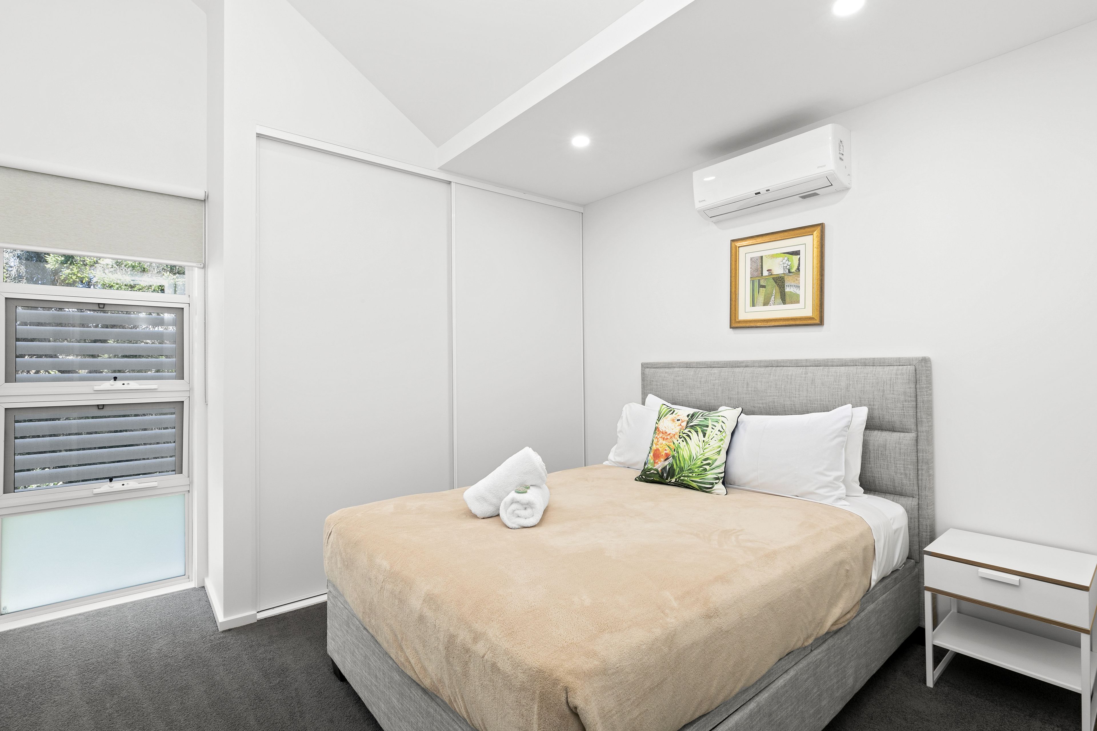A bedroom with a bed, a nightstand, a window with blinds, and an air conditioning unit.