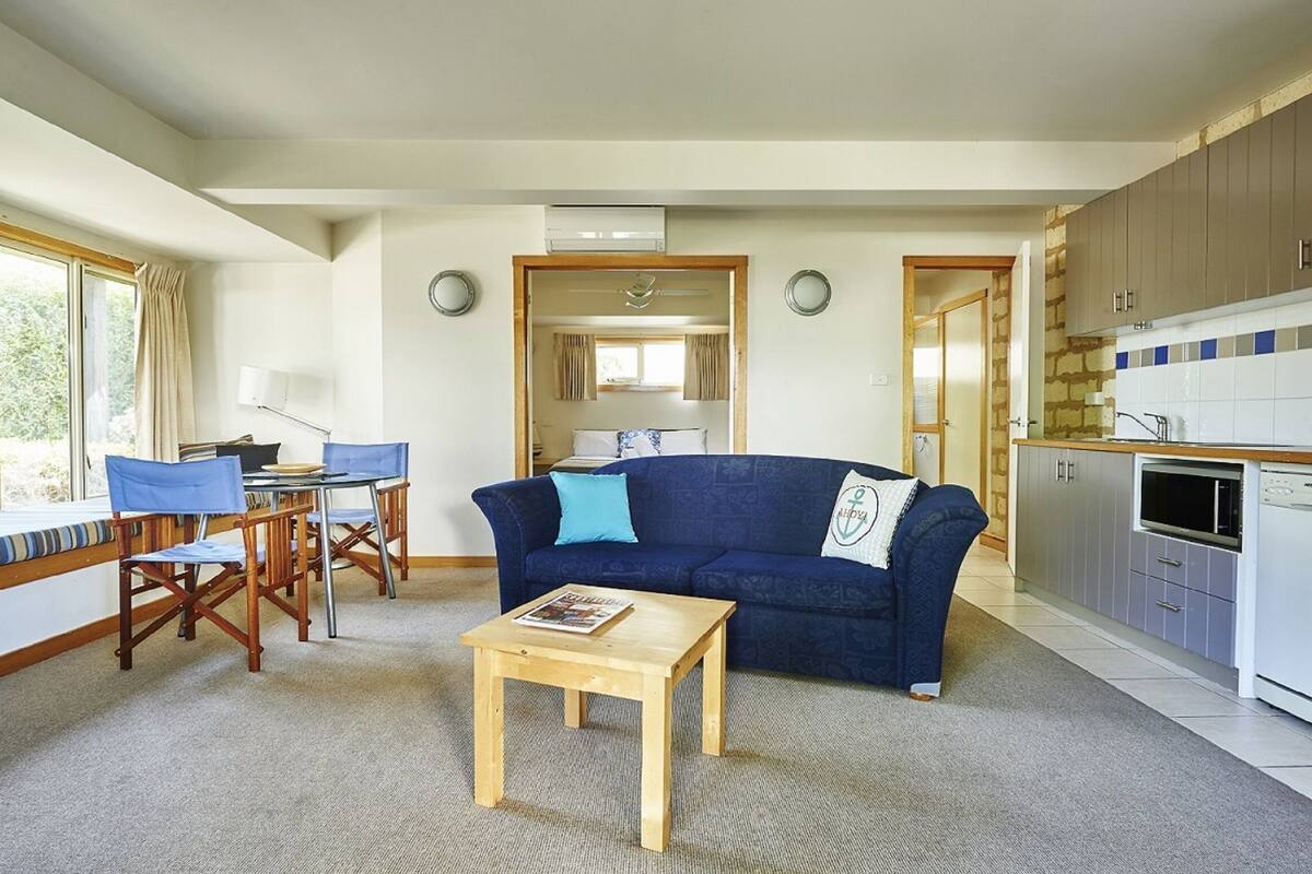 A compact living space with a sofa, dining table, and chairs, a kitchenette, and a view through an open door.