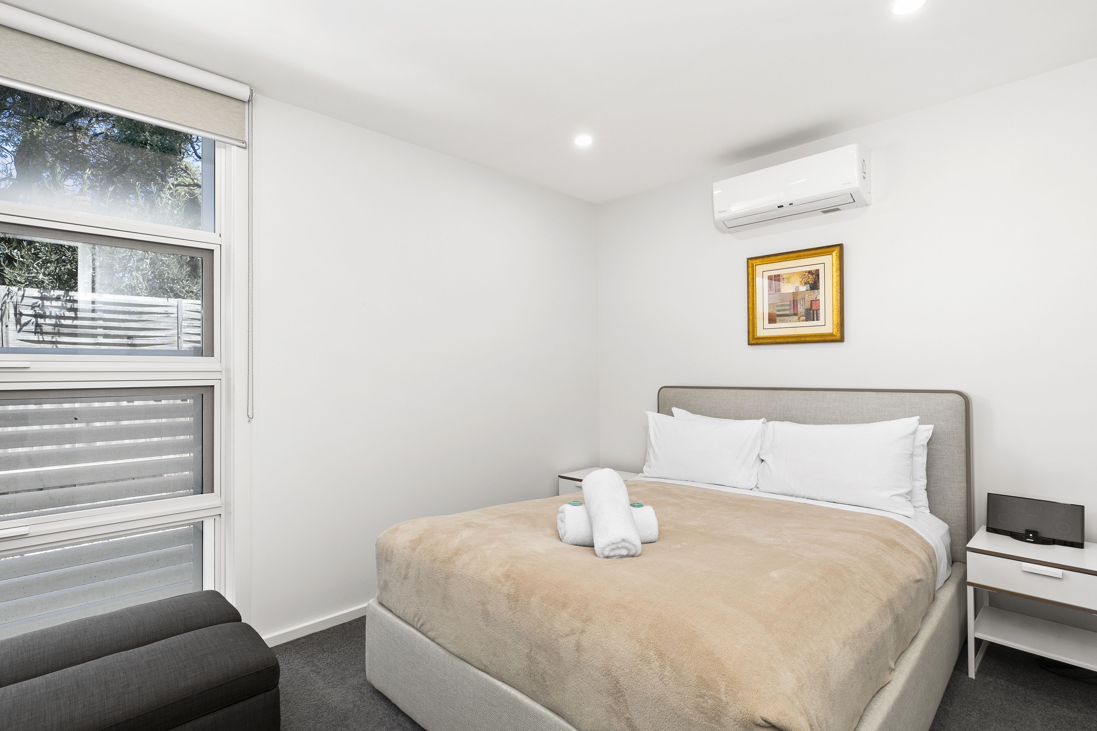 Une chambre d’hôtel comprenant un lit, une table de chevet, un climatiseur, une fenêtre avec des stores et un tableau encadré au mur.
