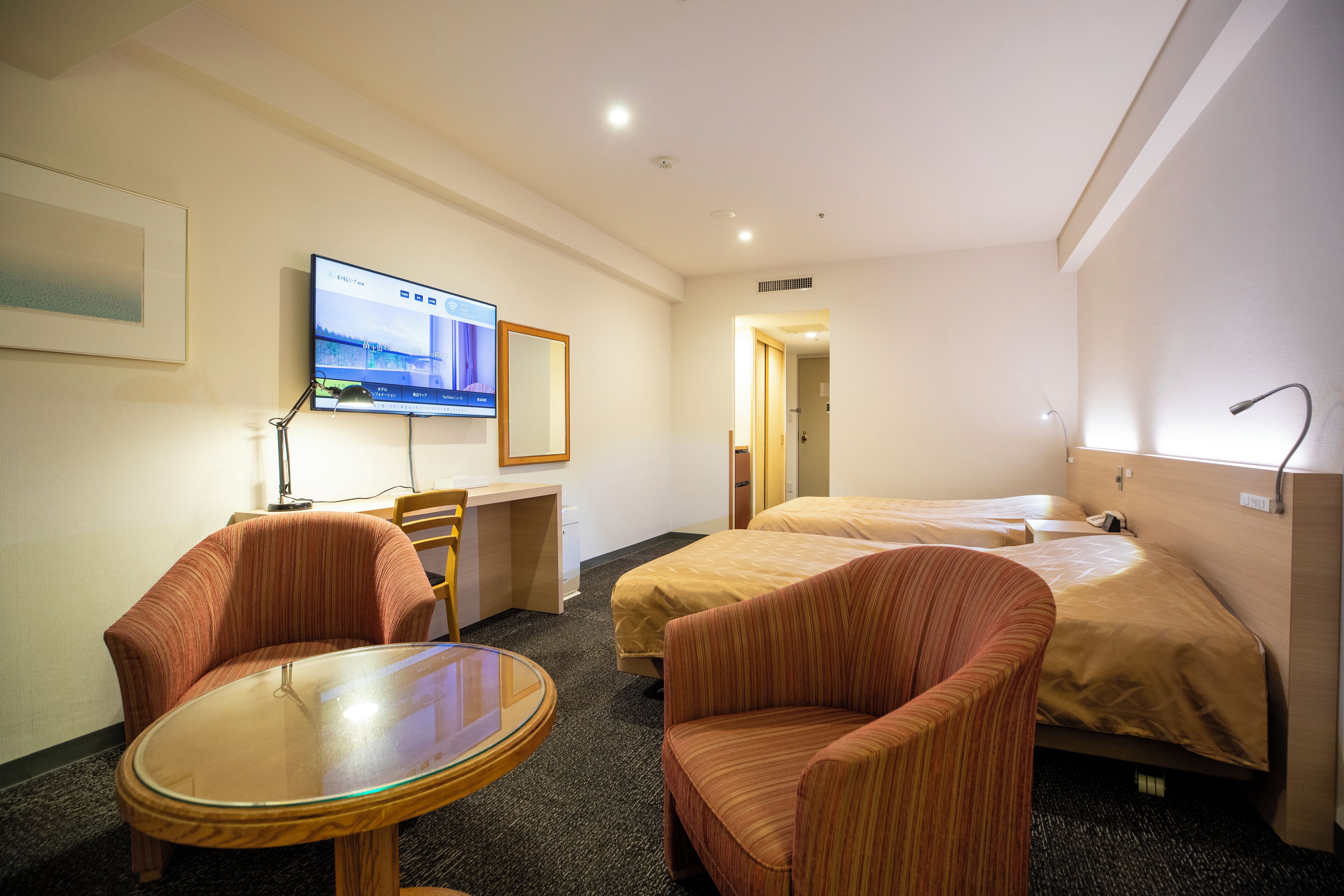 A hotel room with two beds, a desk, a chair, a TV, and a door leading to another room.