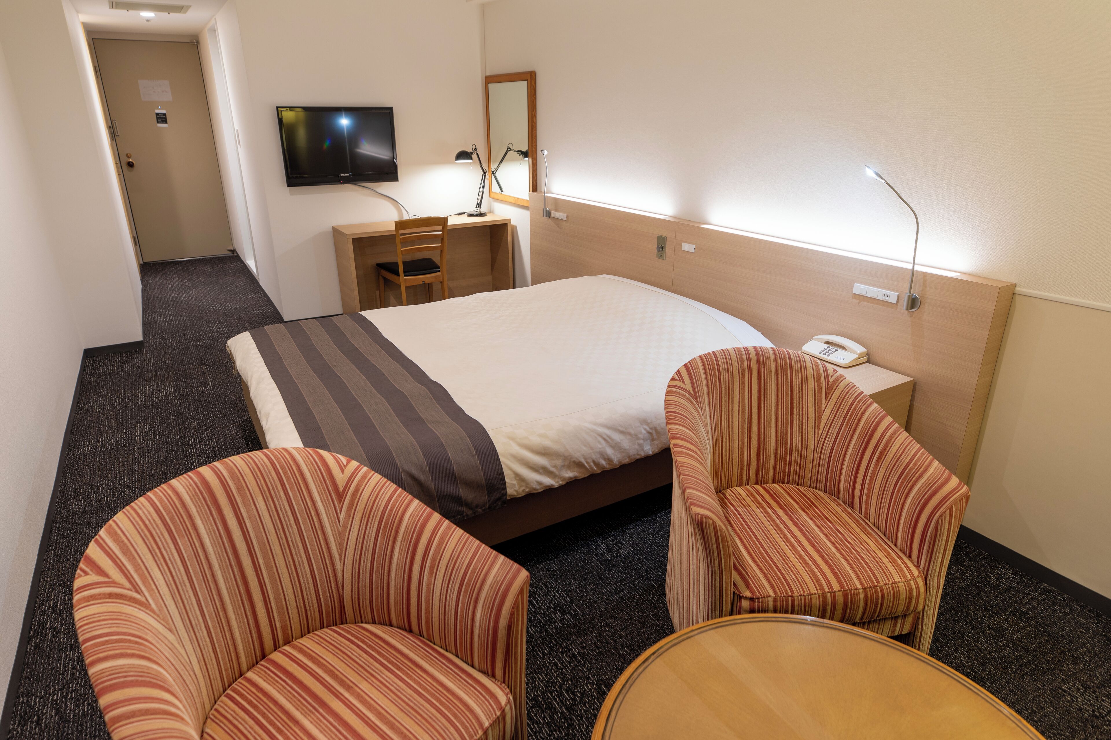 Ein Hotelzimmer mit einem großen Bett, zwei gestreiften Sesseln, einem Schreibtisch mit Stuhl, einem an der Wand befestigten Fernseher und einem Nachttischtelefon.