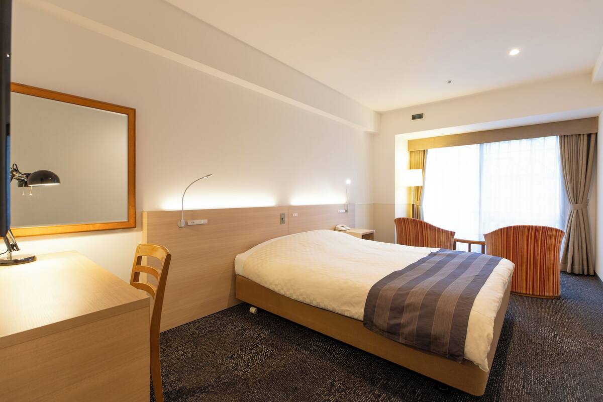 Habitación de hotel con cama, escritorio, silla y un amplio ventanal con cortinas.