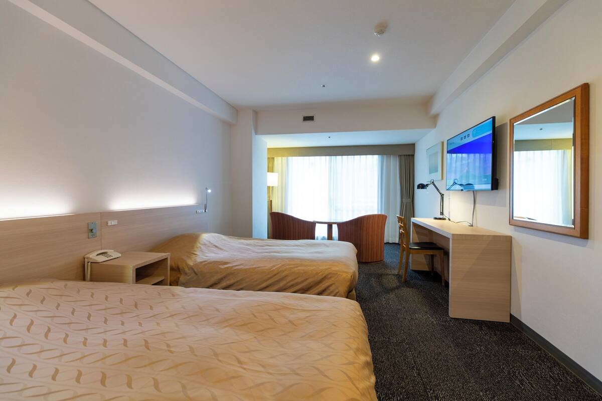 Una habitación de hotel con dos camas, un escritorio, una silla, un televisor y una ventana con cortinas.