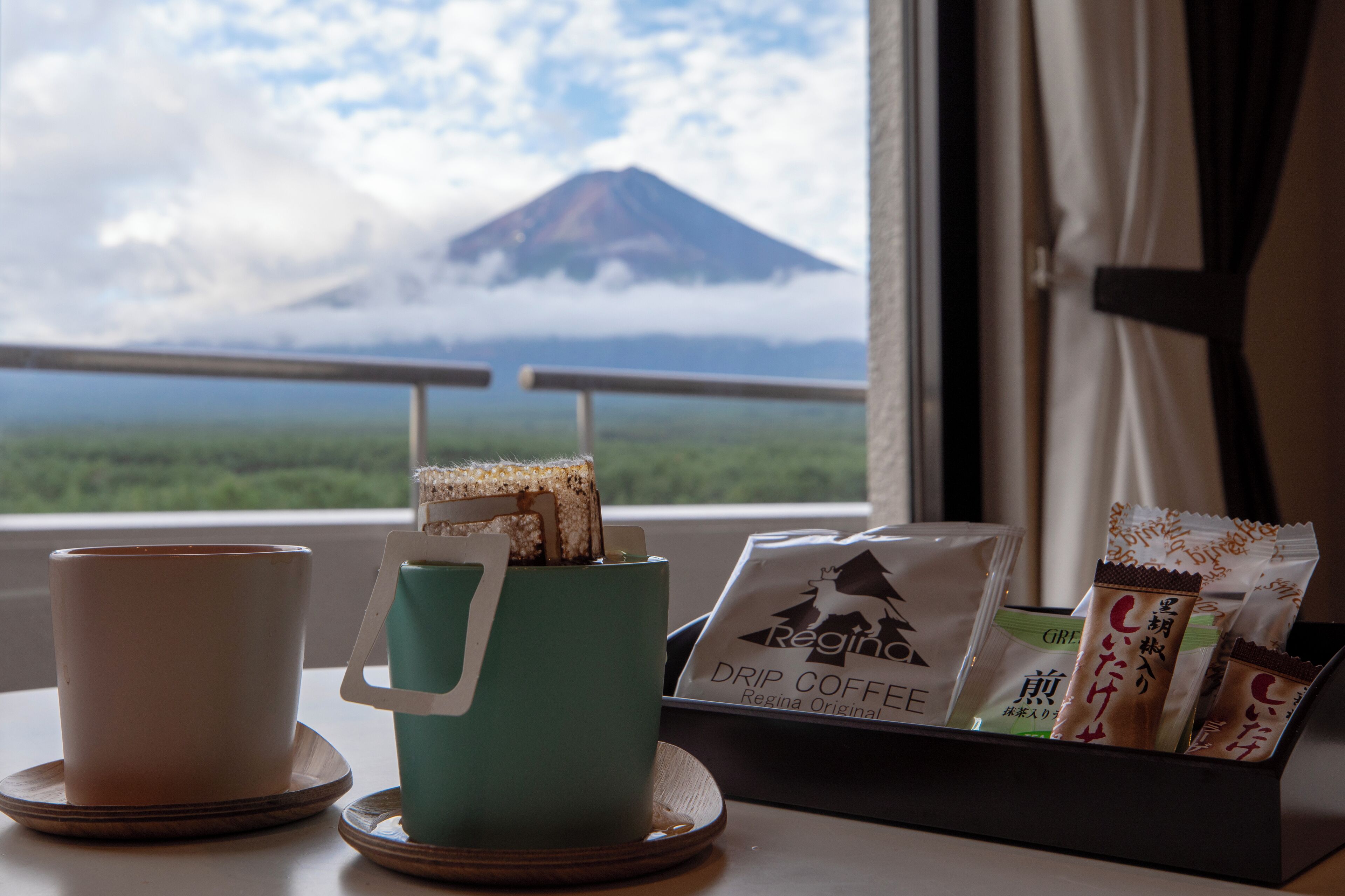 A window with a view of a mountain and clouds, a teal mug with a handle, a white cup, and a tray with coffee packets.