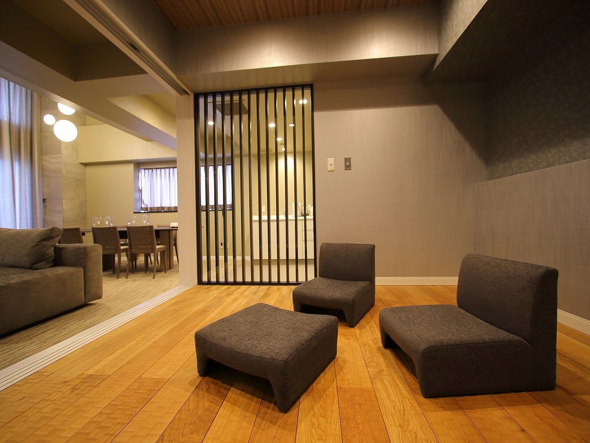 A modern interior with a seating area featuring dark grey sofas and ottomans, wooden flooring, and a partition with vertical slats.