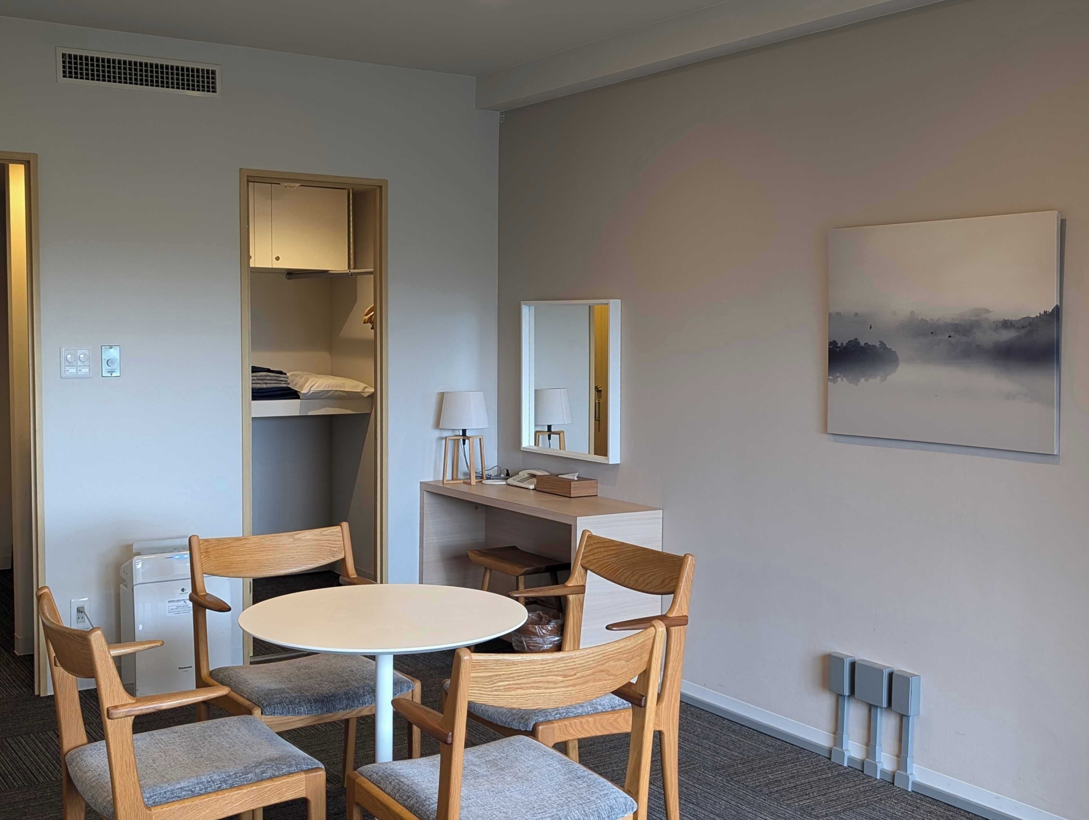 A hotel room with a bunk bed, a desk with a lamp, a chair, a round table, a mirror, and a painting on the wall.
