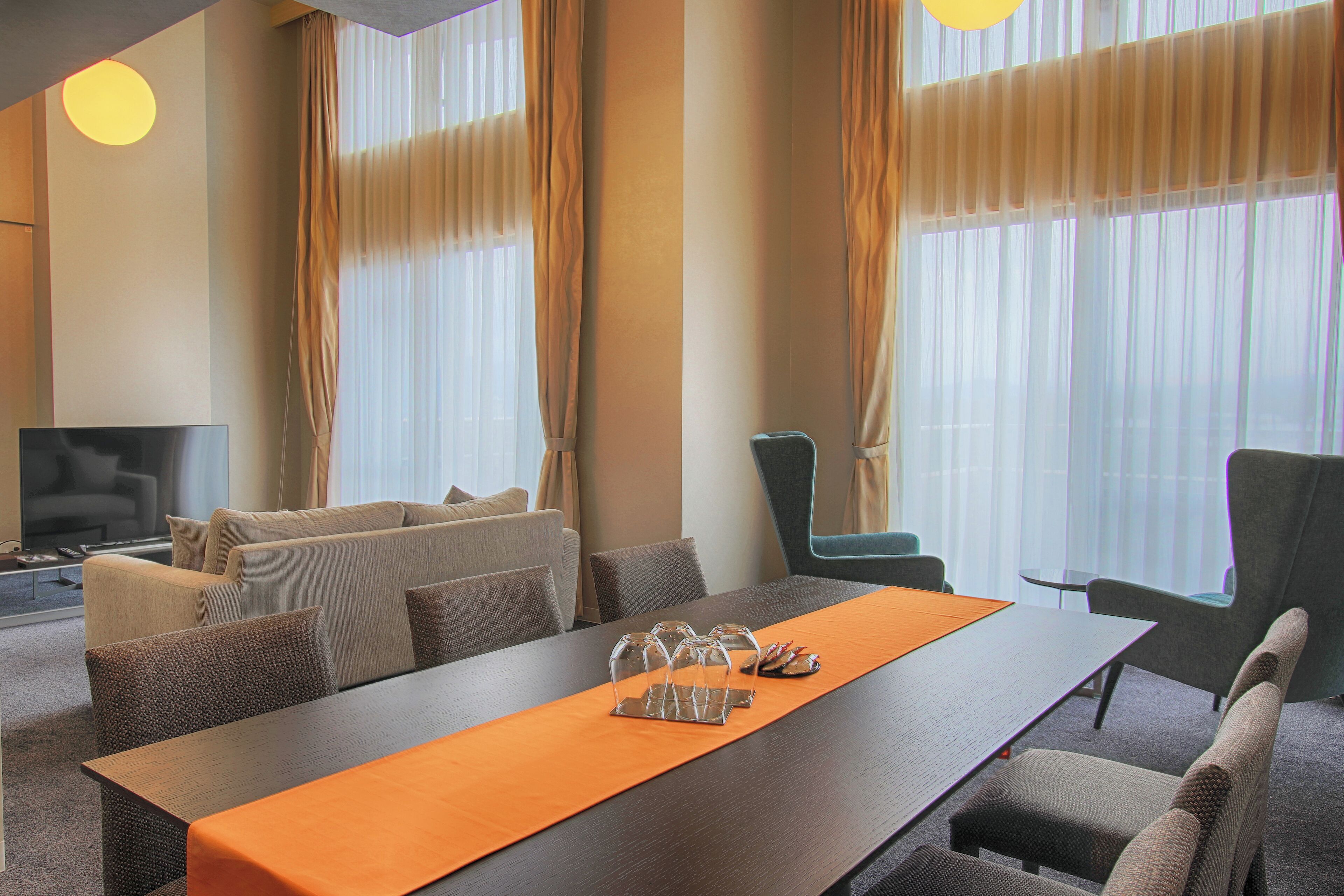 Une chambre d’hôtel moderne dotée d’une longue table à manger, de sièges confortables, d’un téléviseur à écran plat et de grandes fenêtres avec des rideaux.