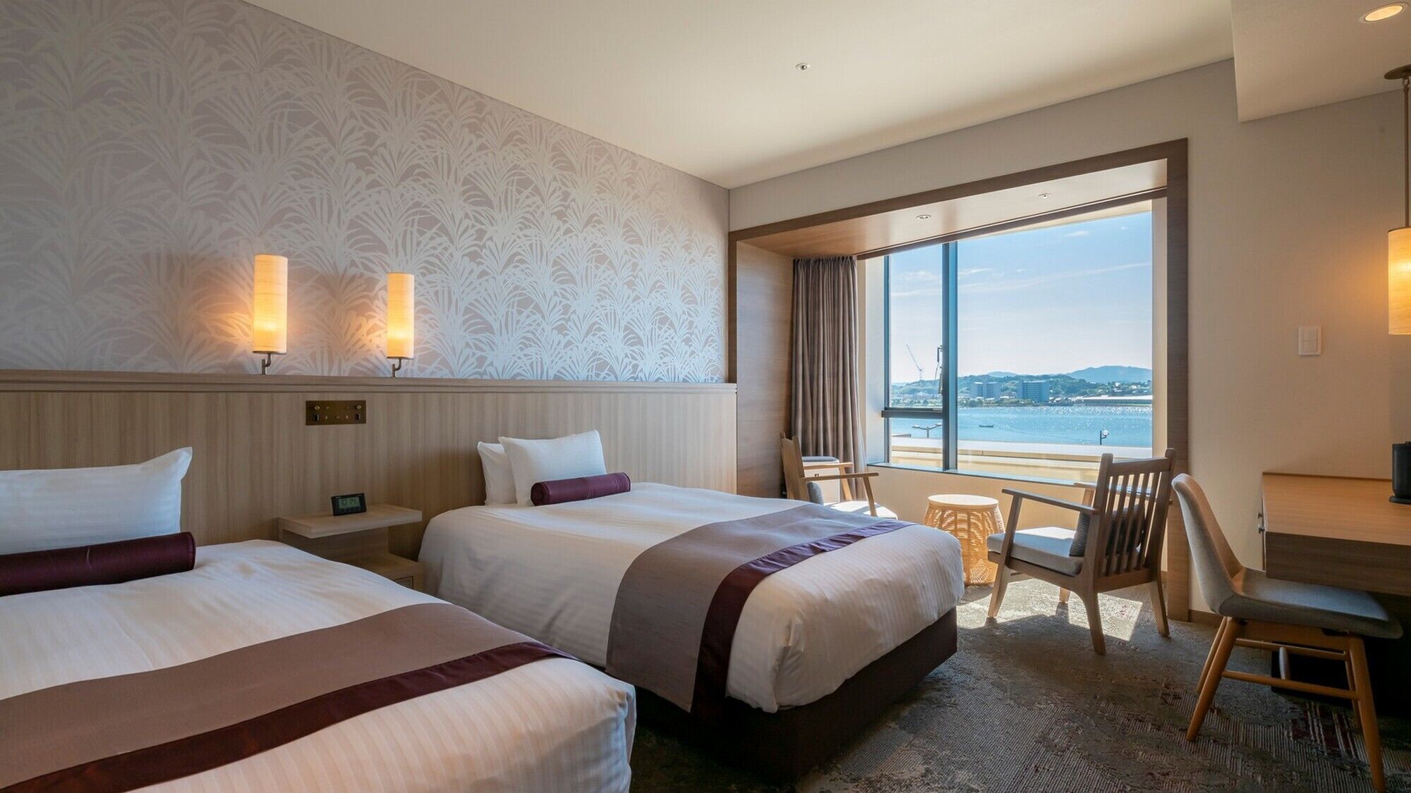 A hotel room with two beds, a desk, a chair, and a view of the sea.