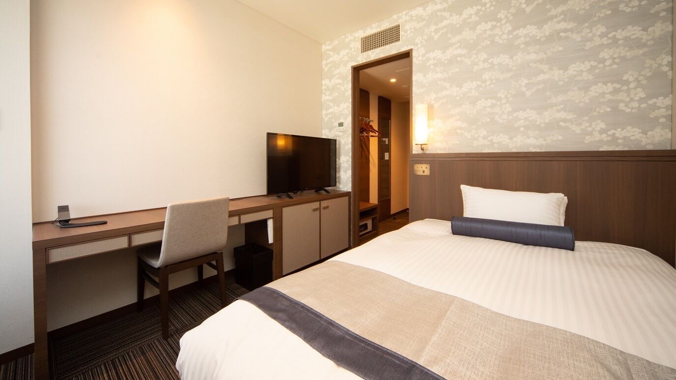 A hotel room with a bed, a desk with a television, a chair, and a wardrobe.