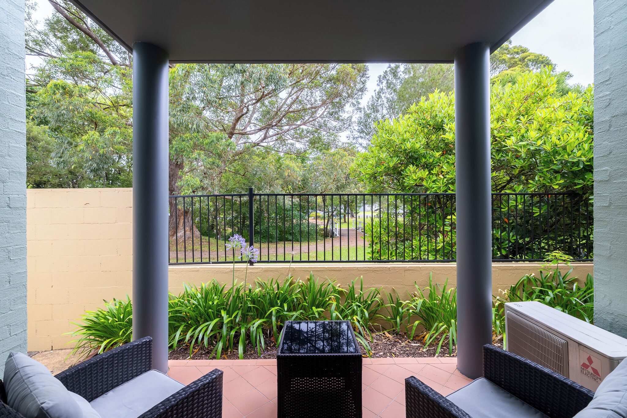 A covered patio area with two wicker chairs and a small table, overlooking a park with a fence and greenery.