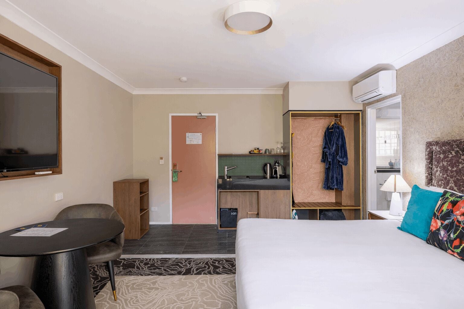 A hotel room with a bed, a desk, a chair, a TV, a kitchenette, and a bathroom.