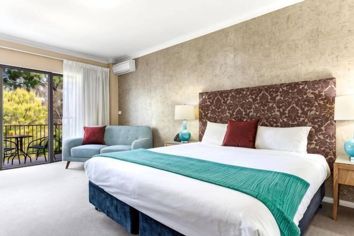 A hotel room with a large bed, a balcony, a chair, and a nightstand with a lamp.