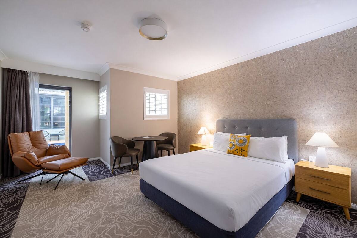 A hotel room with a bed, a chair, a table, and a lamp.