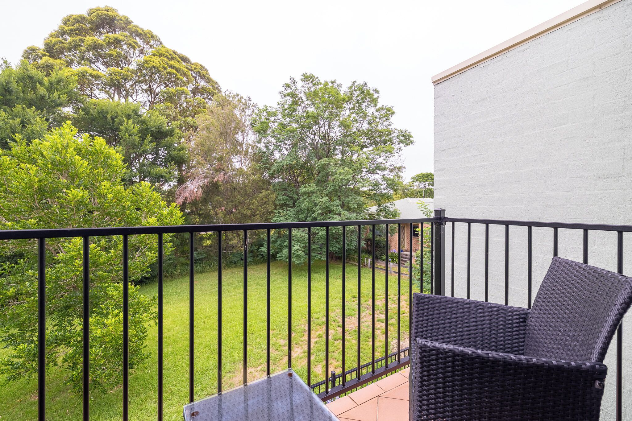 A balcony with a wicker chair and a small table, overlooking a grassy yard with trees.