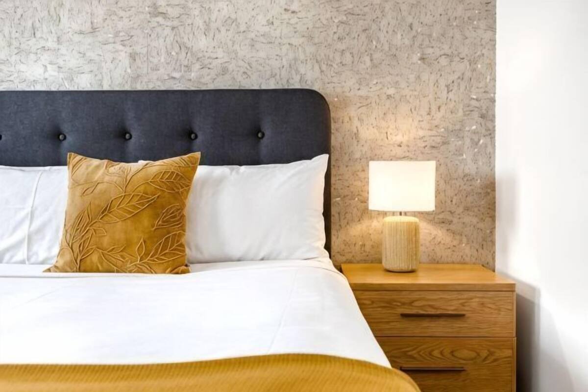 A bed with a dark headboard, white bedding, and a single decorative pillow. A bedside table with a lamp is visible.
