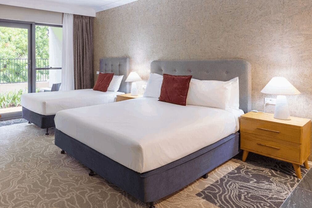 A hotel room with two beds, a grey headboard, white bedding, a patterned rug, and a wooden nightstand with a lamp.