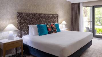 A hotel room with a large bed, two bedside lamps, a patterned headboard, and a view of greenery through a window.
