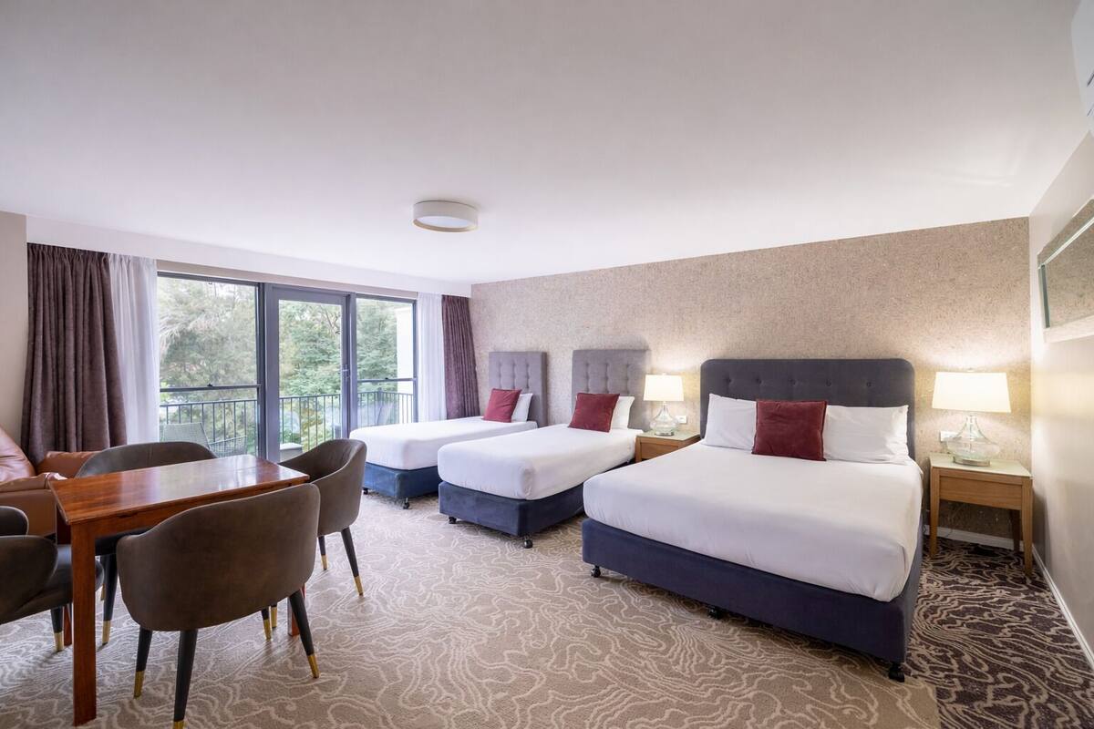 A hotel room with two beds, a dining table, chairs, and a balcony with a view.
