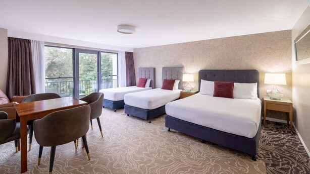 A hotel room with two beds, a dining table, chairs, and a balcony with a view.