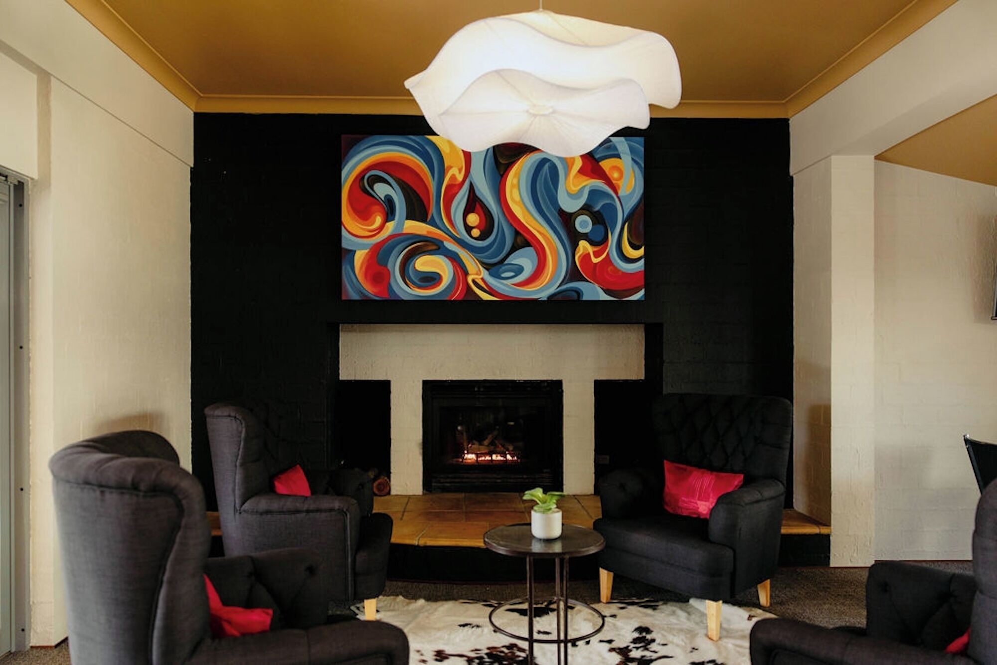 Uma sala de estar moderna com lareira, sofás pretos com almofadas vermelhas, uma mesa de centro redonda e uma pintura abstrata colorida acima da lareira.