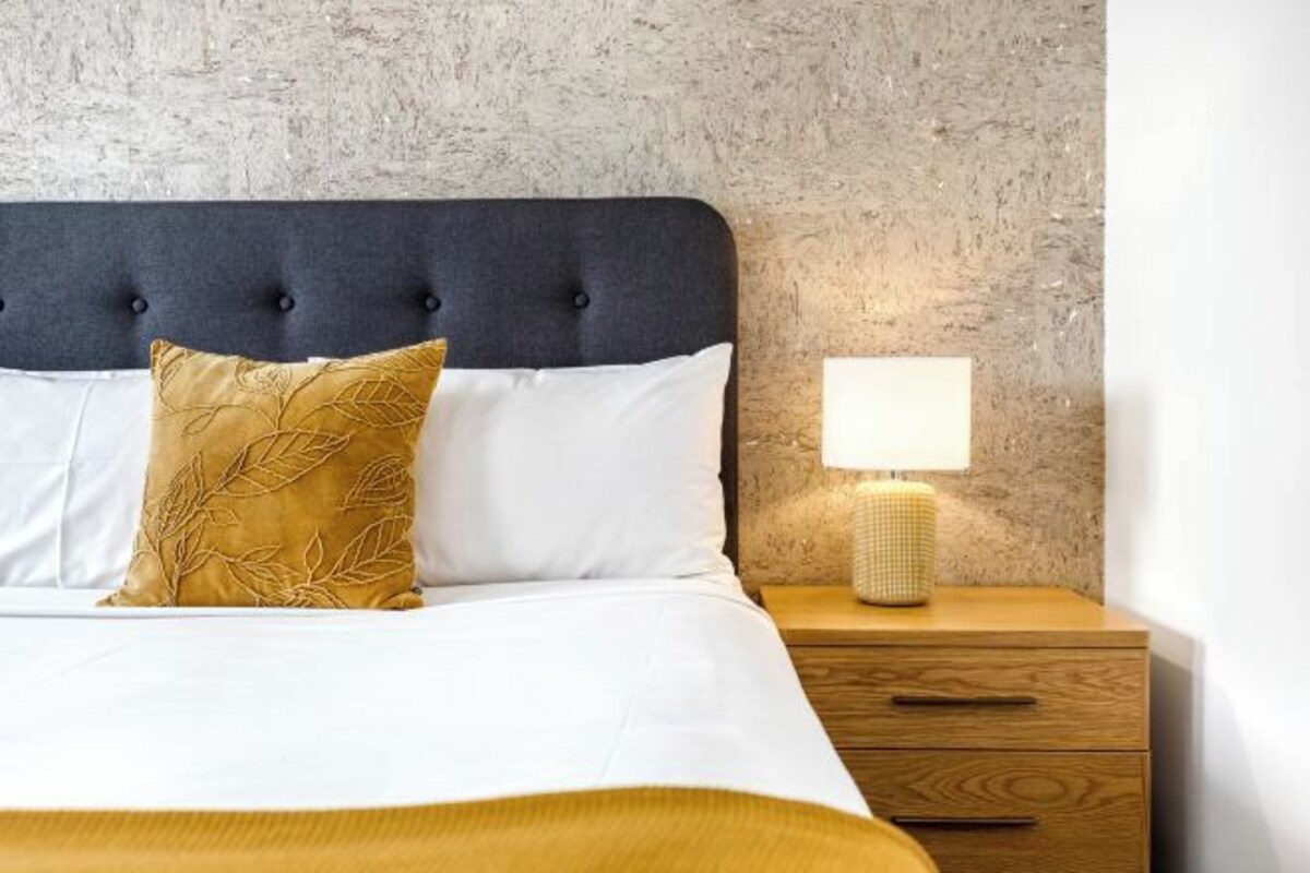 A neatly made bed with a dark headboard, a white pillow, a gold-patterned pillow, and a white bedsheet. There is a wooden nightstand with a lamp and a drawer.