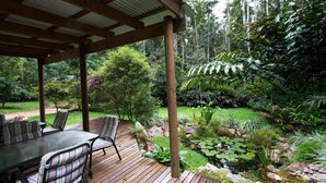 Un espacio al aire libre cubierto con una terraza de madera, asientos y un jardín con rocas, plantas y un estanque.