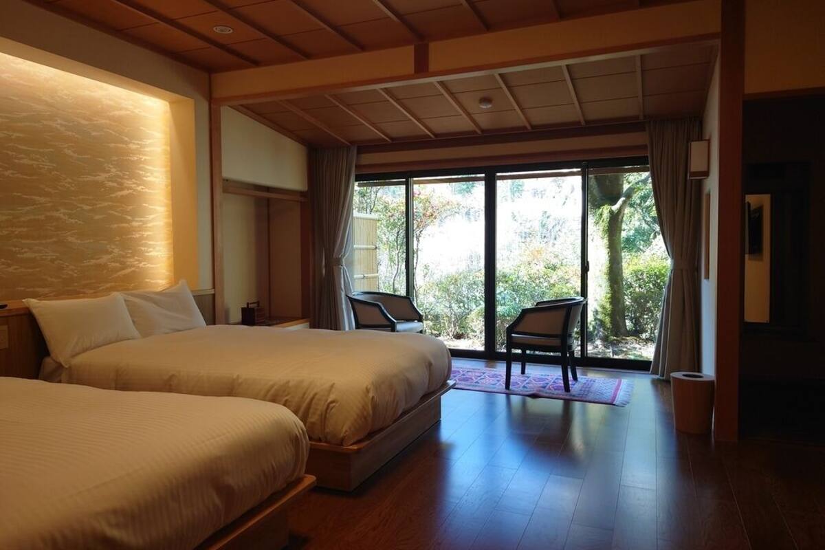 A hotel room with two beds, a large window, and a view of greenery outside.