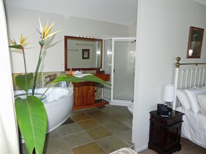 Un baño con bañera, ducha, lavabo e inodoro. Hay una planta grande en la esquina y una mesita de noche con lámpara.
