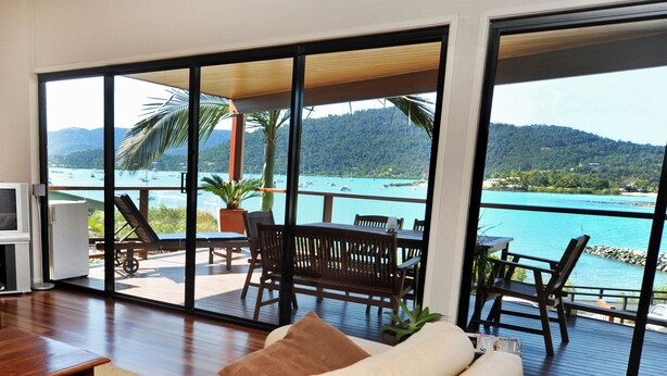 Een ruime kamer met een grote schuifglazen deur die uitkomt op een terras met uitzicht op een turquoise zee en boten.