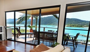Amplia habitación con un gran ventanal corredizo que da a una terraza exterior con vistas al mar turquesa y embarcaciones.