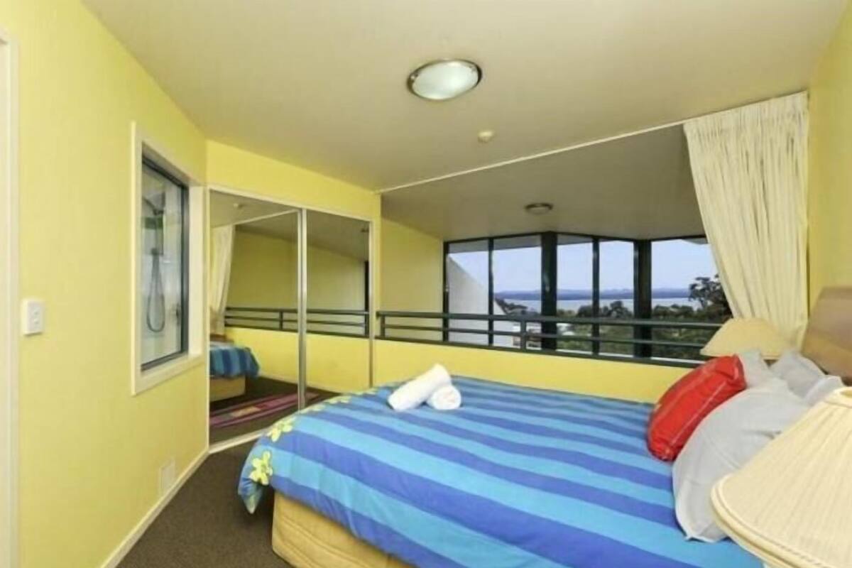 A hotel room with a bed, a window with a view, a mirror, and a shower.