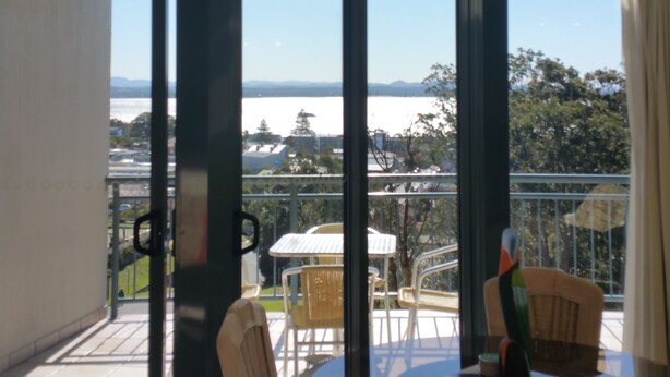 A room with large windows offering a view of a body of water and distant mountains. There are two chairs and a table in the foreground. - Bali in Broughton (Nelson Bay)
