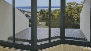 A corner room with large windows offering a view of a lake, buildings, and trees. - Bali in Broughton (Nelson Bay)