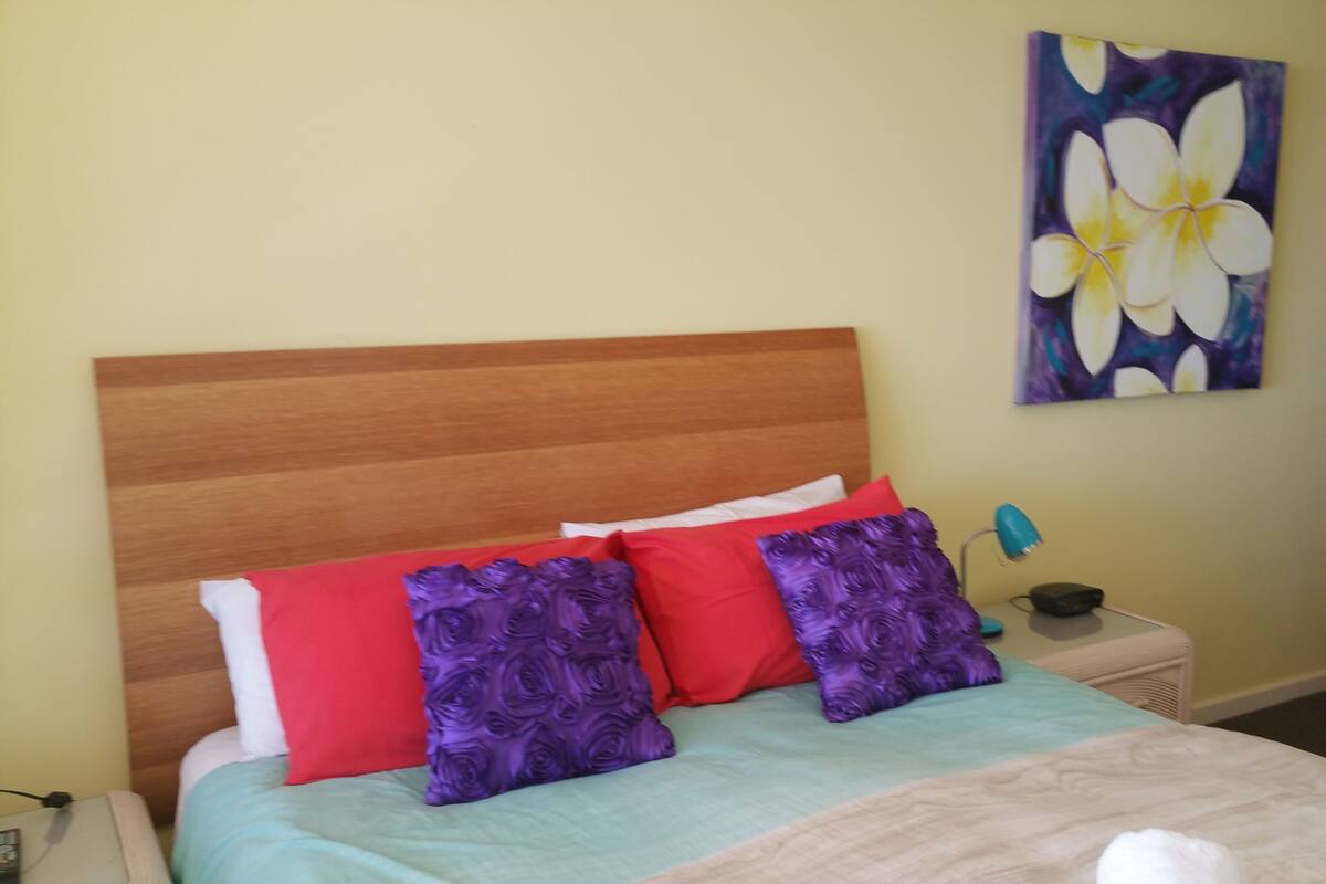 A bed with a wooden headboard, red and purple pillows, a teal bedspread, and a white blanket. There is a painting of flowers on the wall above the bed.