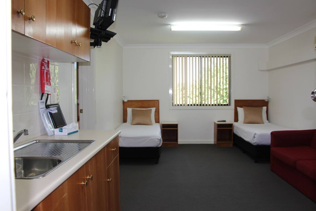 A hotel room with a kitchenette, two beds, a red sofa, and a window with blinds.