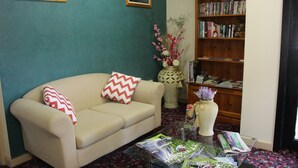 A living room with a beige sofa, two pillows, a glass coffee table with magazines, a bookshelf, and a vase with flowers. - Pioneer Way Motel (Faulconbridge)