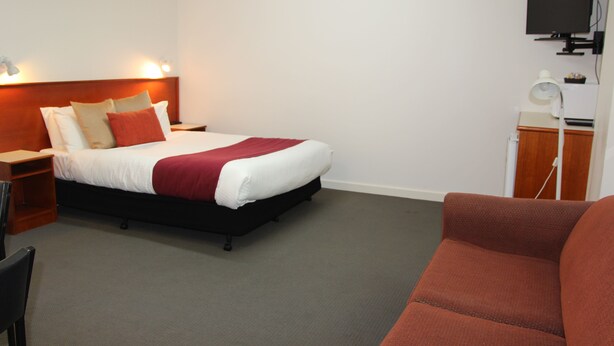 A hotel room with a bed, a desk, a chair, a TV, and a red sofa. - Pioneer Way Motel (Faulconbridge)