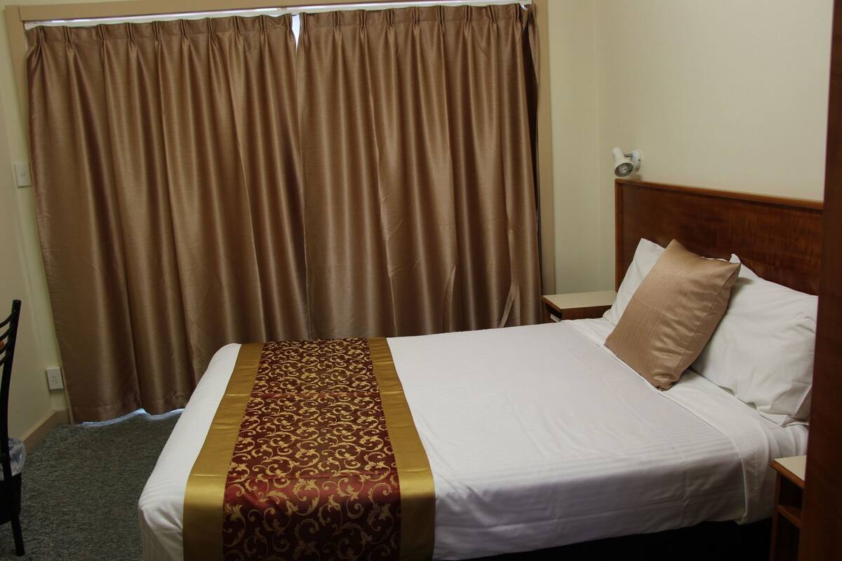 A hotel room with a bed, a nightstand, a lamp, a chair, and a curtain.