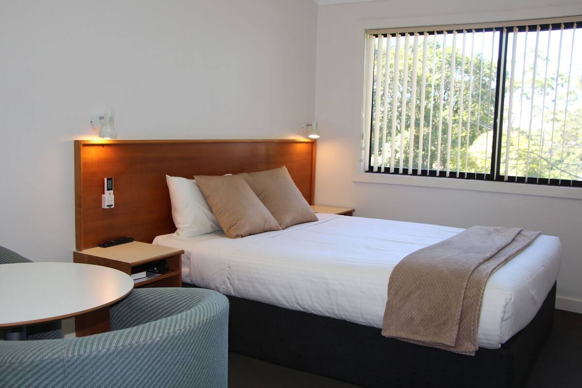 A hotel room with a bed, bedside tables, a desk, and a window with blinds.