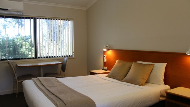 A hotel room with a bed, bedside table, lamp, and a desk with a chair. - Pioneer Way Motel (Faulconbridge)