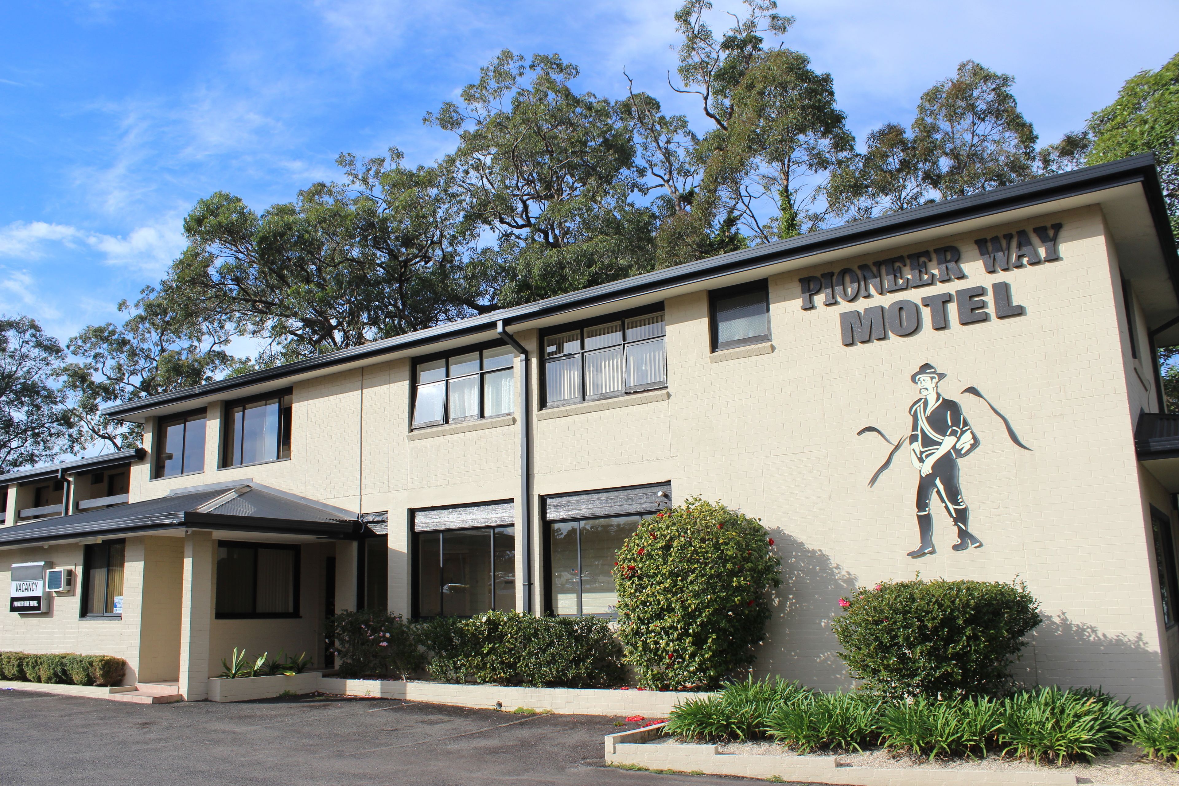 A Pioneer Way Motel with a large mural of a cowboy on the side, a parking lot, and surrounding greenery.