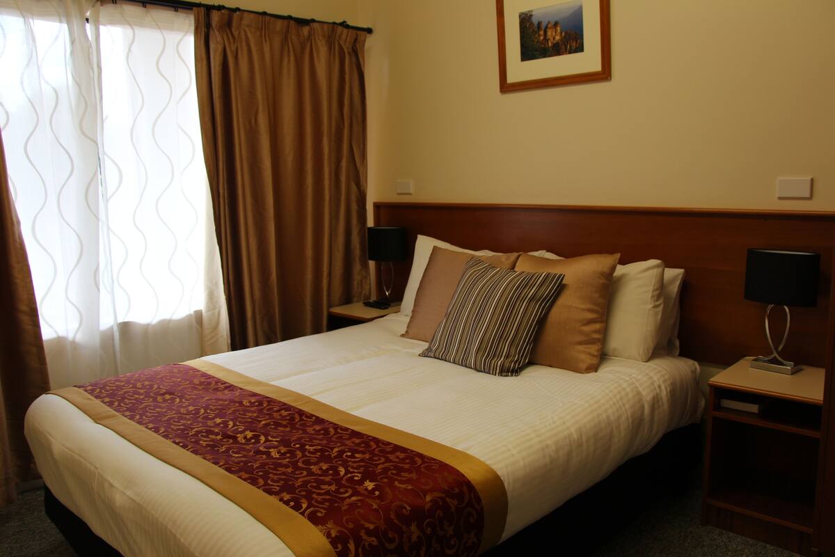 A hotel room with a large bed, two bedside tables, a nightstand, a framed picture on the wall, and a window with curtains.