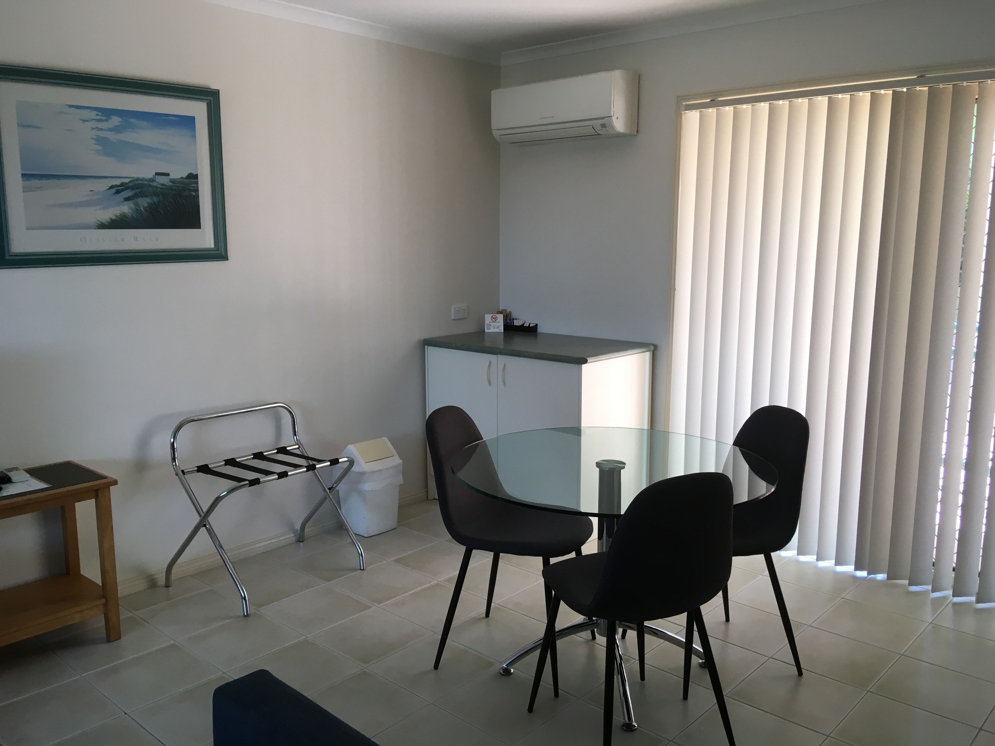A small, clean room with a glass table, two chairs, a metal drying rack, a cabinet, and a window with blinds.