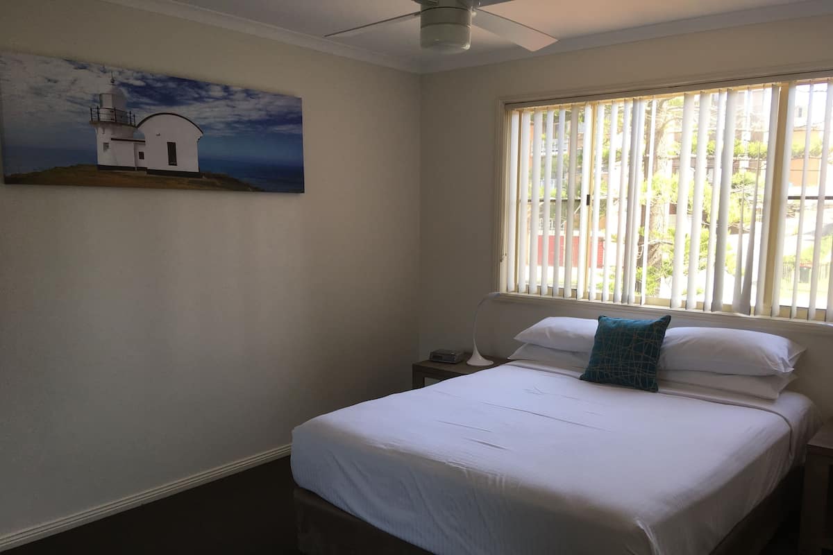 A bedroom with a bed, a nightstand, a lamp, a painting of a lighthouse, and a window with blinds.