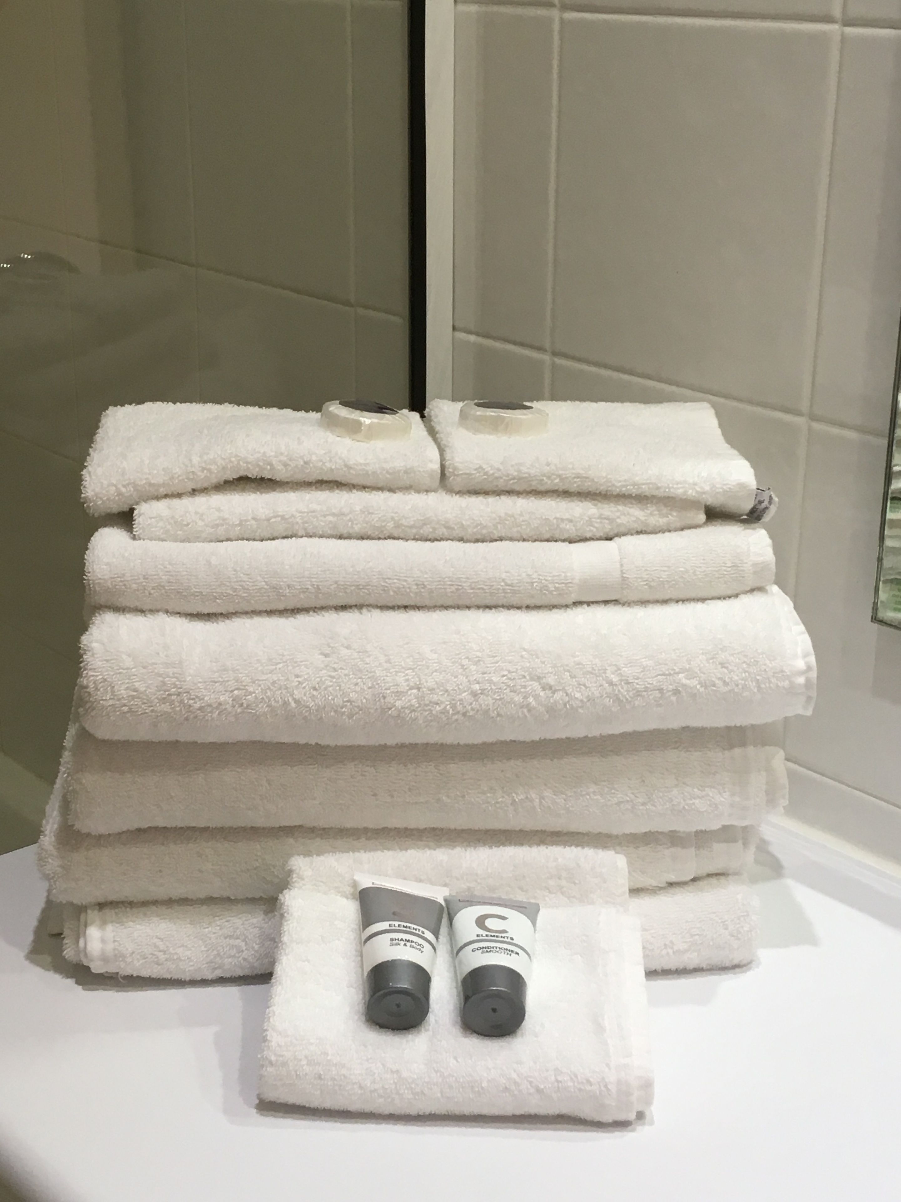 A stack of white towels and two bottles of Elements shampoo and conditioner on a white surface.