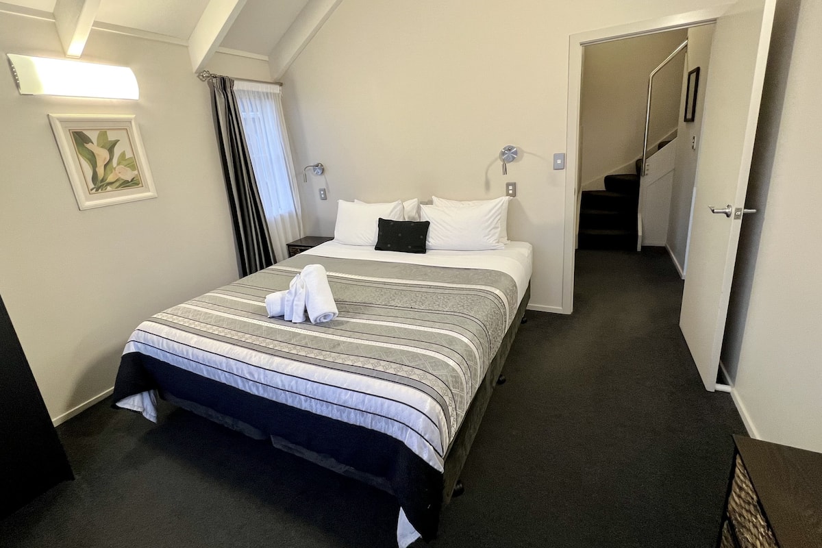 A hotel room with a bed, a nightstand, a framed picture on the wall, and a staircase leading to another room.