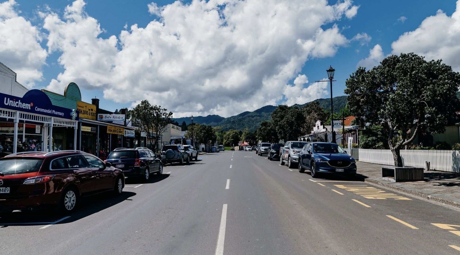 A street lined with parked cars and various shops, including a pharmacy and a restaurant.