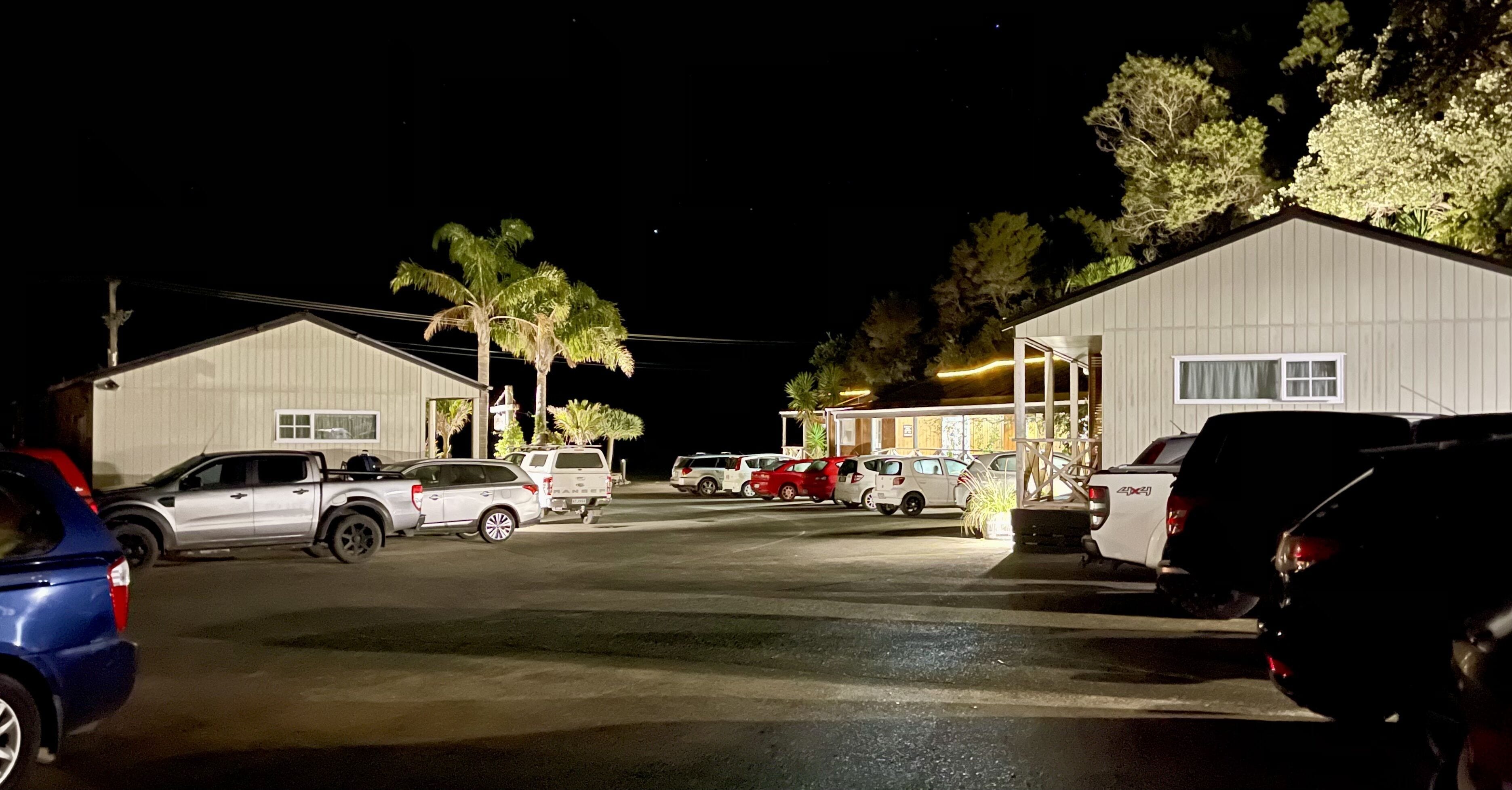 Un estacionamiento con varios vehículos, incluyendo automóviles y un camión, frente a edificios con grandes ventanales y una zona techada.