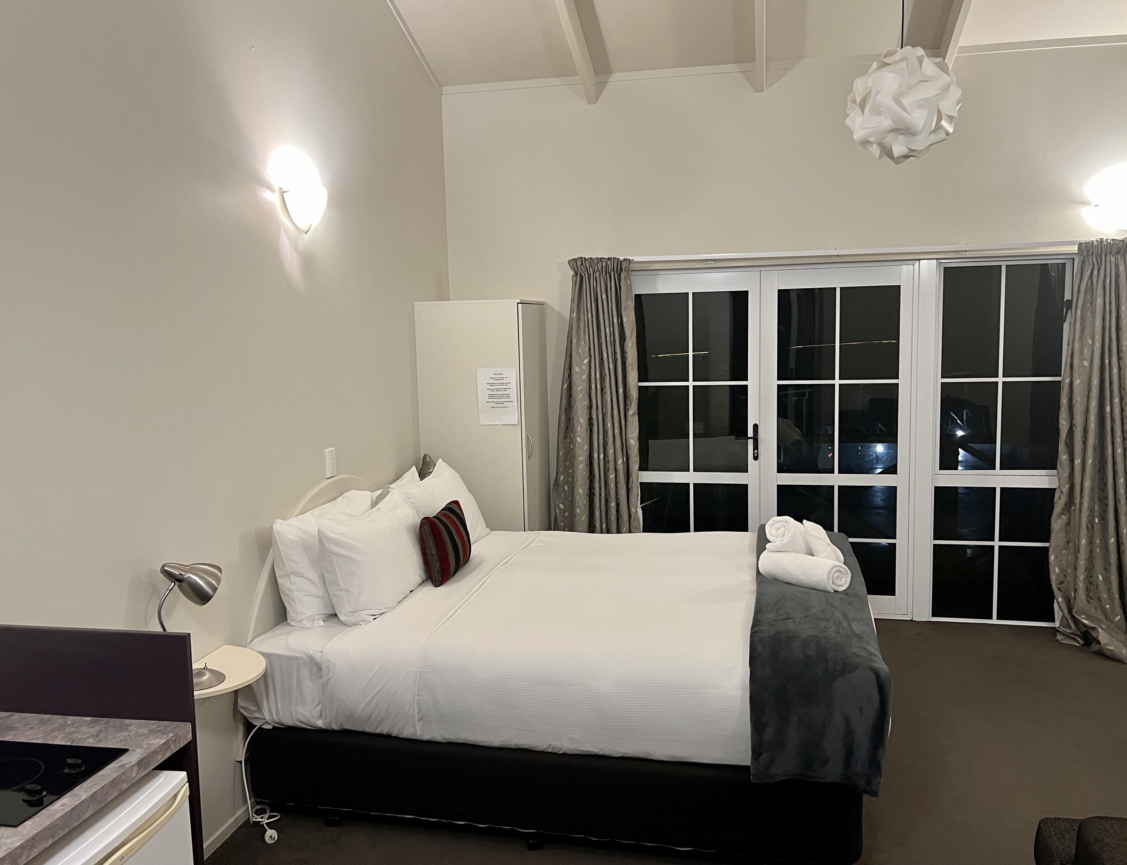 A hotel room with a bed, a nightstand, a desk with a lamp, a window with curtains, and a view of the outside.